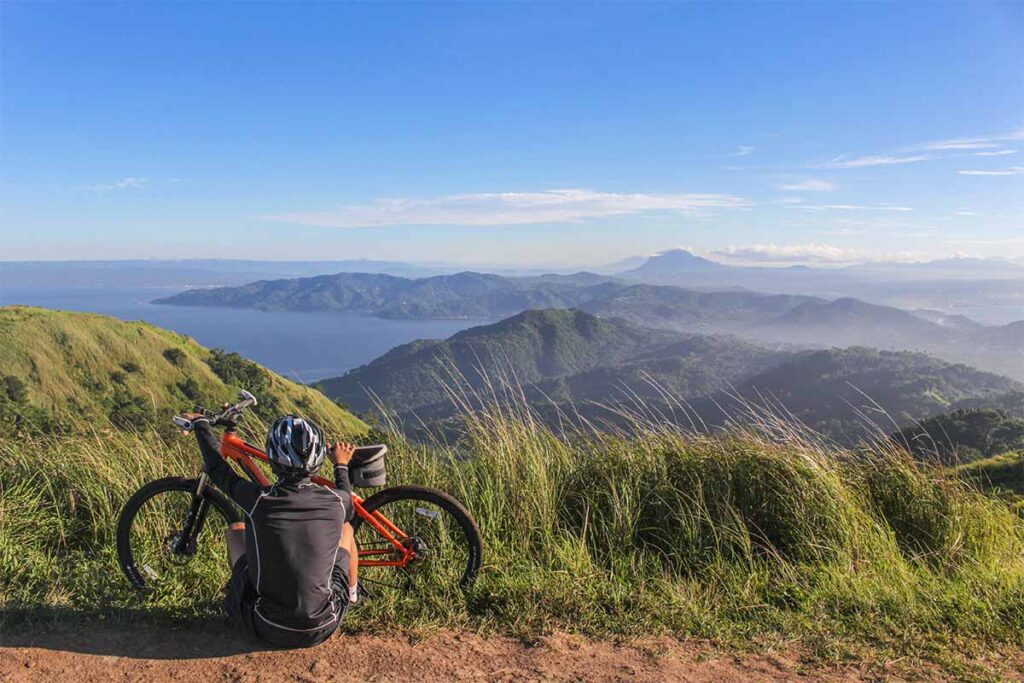 mountain bike - homem sentando no topo da montanha, segurando sua mtb apreciando a paisagem, onde aparecem montanhas e um grande lago de fundo